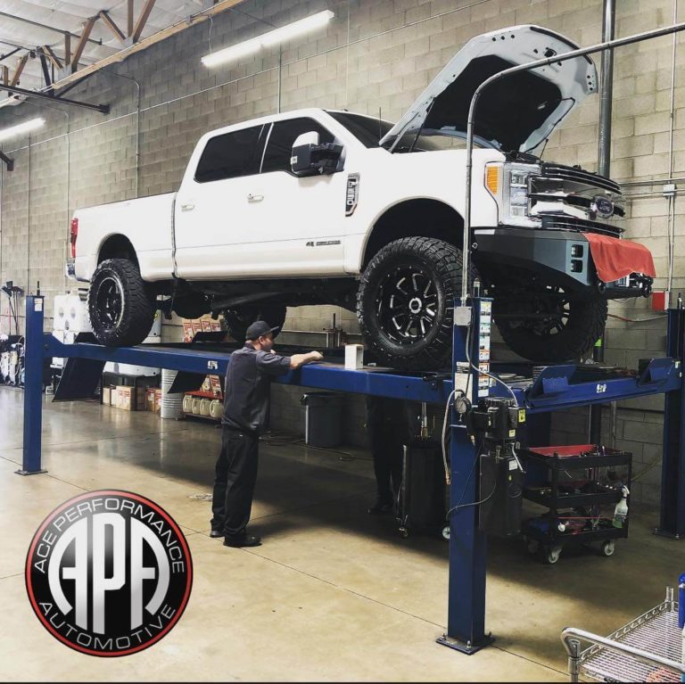 A White Ford F250 on-top of a vehicle lift with the hood raised and a red towel hanging on the front of the bumper
