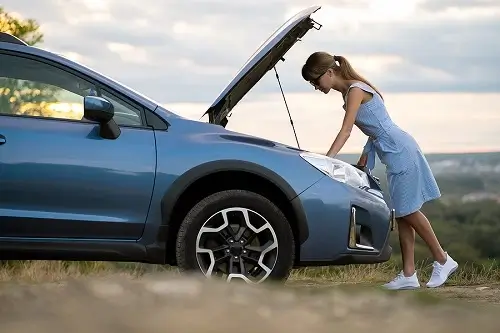 Common Auto Repair Issues Drivers Face in Queen Creek, AZ | Ace Performance Auto. Image of a stranded young woman driver standing near her broken blue car with popped up bonnet, inspecting her car for battery problem or overheating engine.