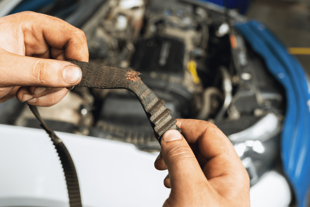 Timing belt replacement in Queen Creek, AZ by APA Auto & Fleet Care. Image showing a mechanic holding a worn, cracked timing belt in front of a vehicle engine, emphasizing the importance of timely belt replacement to prevent costly engine damage.